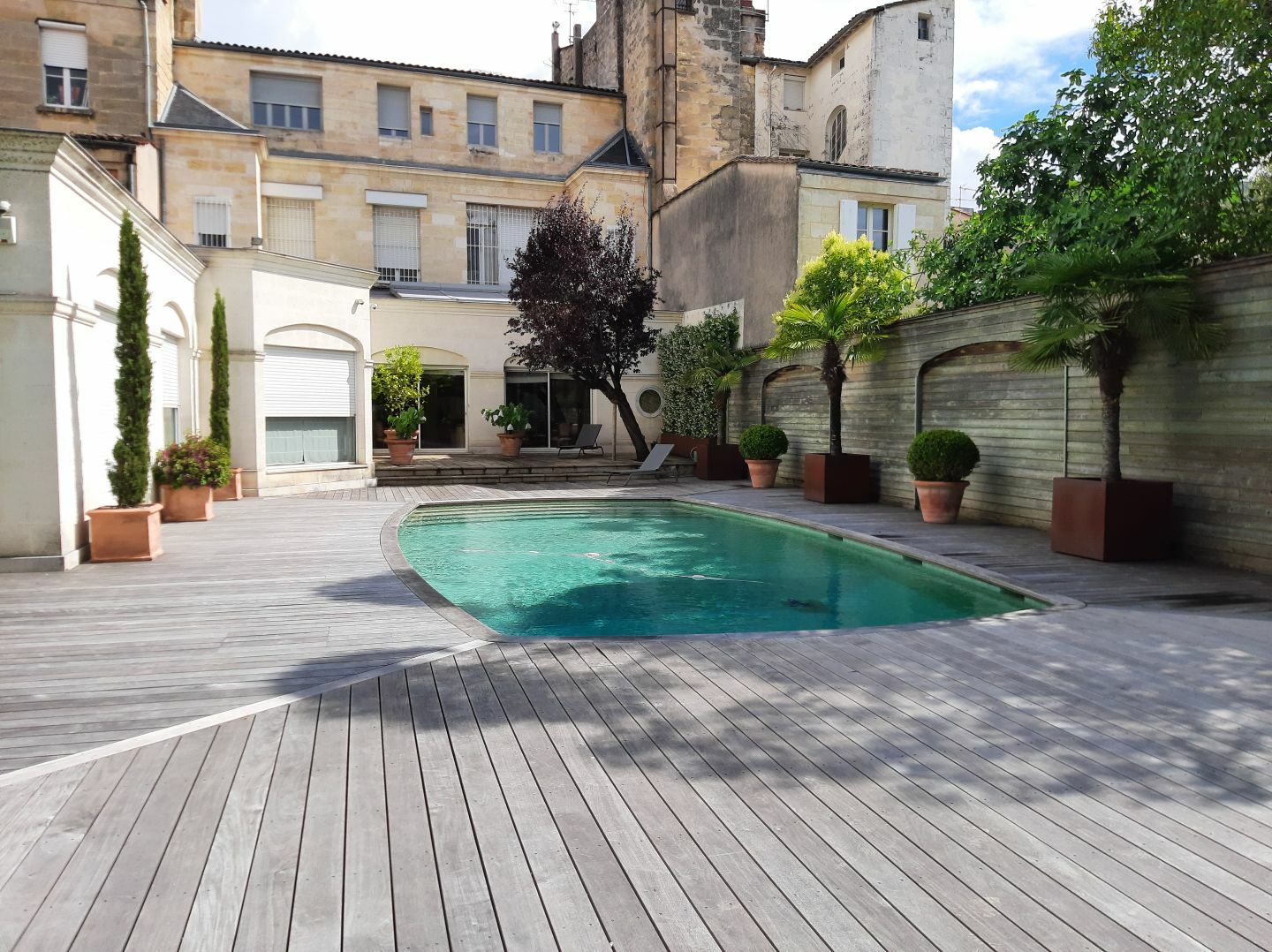 Photo d'un jardin aménagé à Bordeaux avec la réalisation d'une terrasse en bois autour de la piscine. On peut également déceler diverses plantations en pot. 